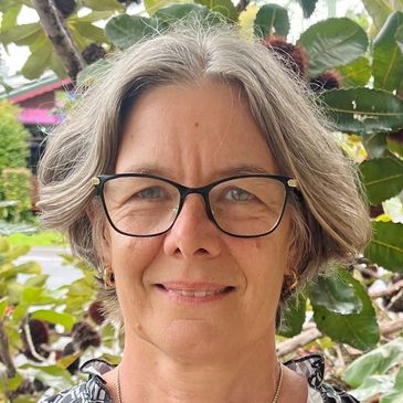 Smiling woman with glasses standing outdoors in front of leafy plants.