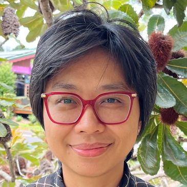 Person with short black hair and red glasses smiling outdoors near leafy trees.