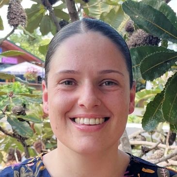 Smiling woman in colorful patterned top stands outdoors near leafy plants.