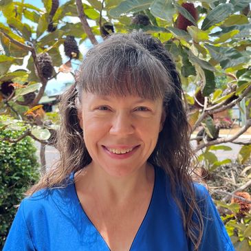 Smiling woman with long hair in blue shirt outdoors near plants.