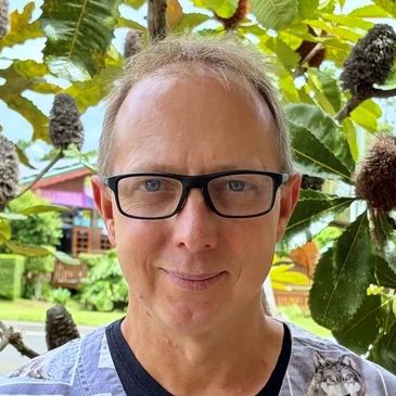 A man with glasses stands outdoors among lush foliage, wearing a shirt with animal prints.