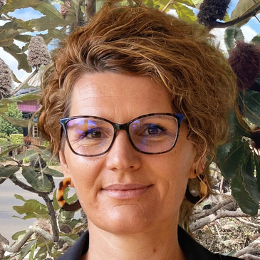Woman with glasses and curly hair standing outdoors near leafy plants.