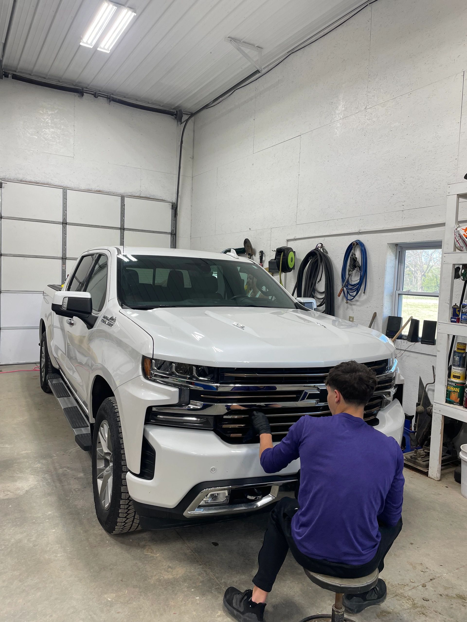 Man working on the front grille of a white Chevrolet truck in a garage.