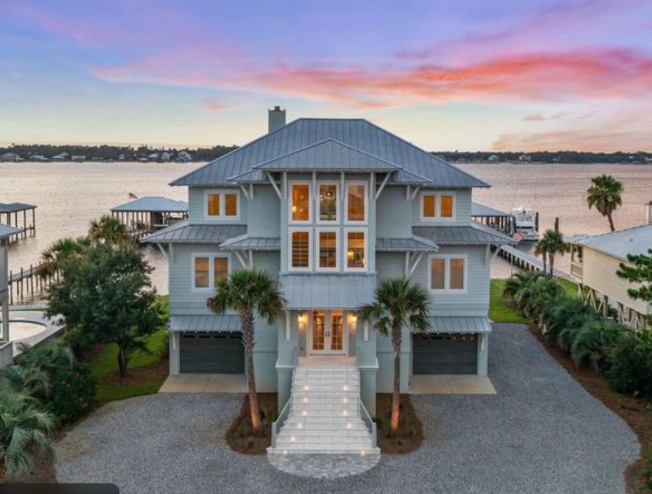 Large waterfront house with a grand staircase and sunset sky.