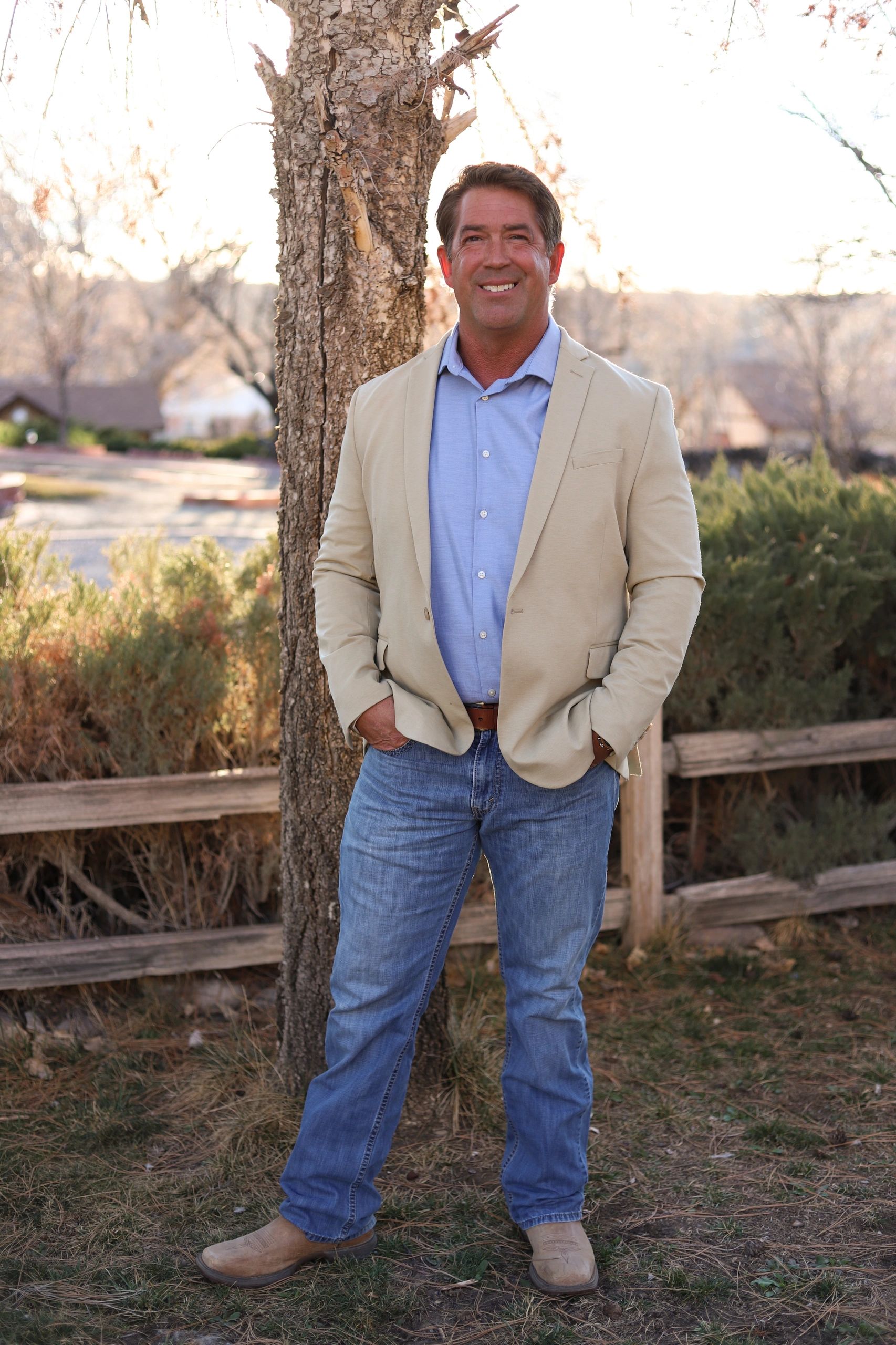 Man in casual blazer, jeans, and boots stands smiling outdoors by a tree.