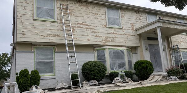 House exterior being prepared for painting with ladders and plastic coverings.