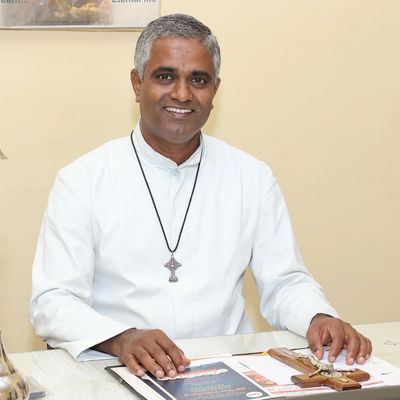 A smiling man in white religious attire sitting at a desk with religious items.