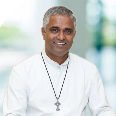 Smiling man in white clerical attire sitting at a table with religious items.