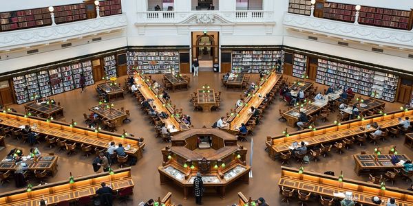 A large, elegant library reading room with wooden desks and green lamps.