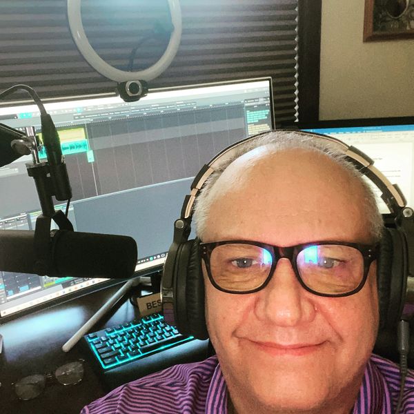 Man with glasses and headphones smiling in front of computer and microphone.
