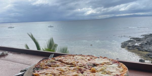 A pizza with cheese and toppings on a wooden plate by the sea.