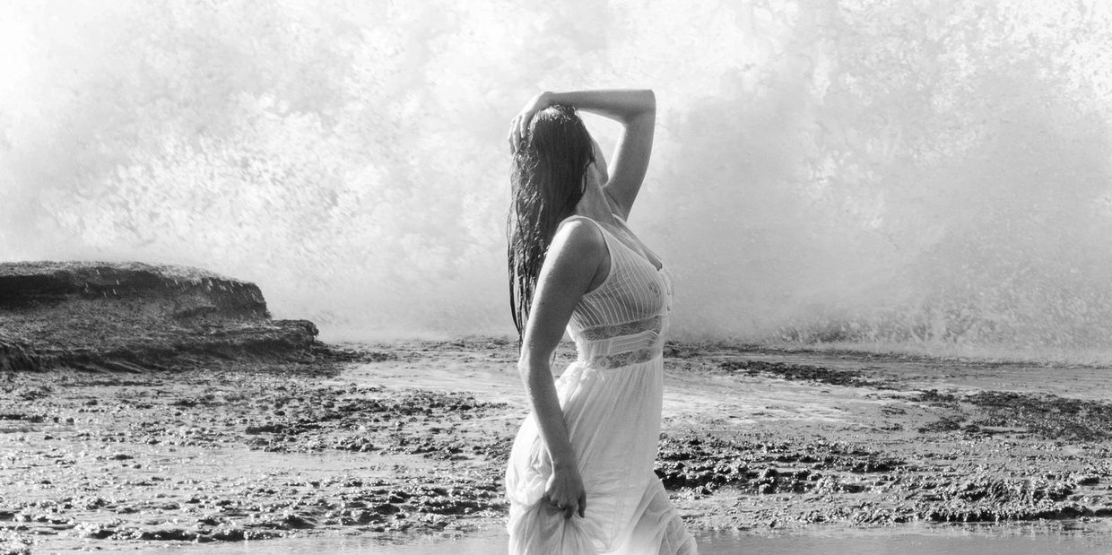 A woman in a white dress poses on a rocky shore with waves crashing behind her.