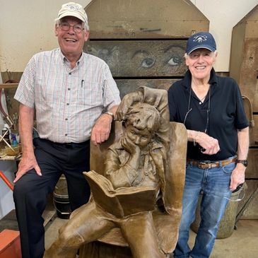 Two elderly men standing beside a bronze sculpture of a boy reading a book.