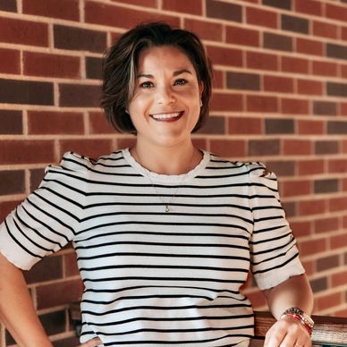 Smiling woman in a striped shirt stands against a brick wall.