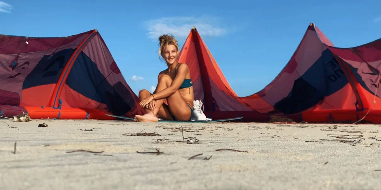 Smiling woman in bikini sitting on sand with colorful kites in background.