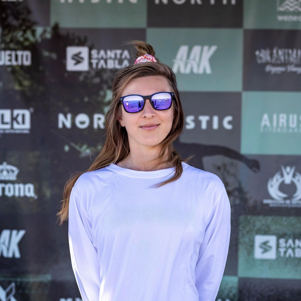 Young woman with sunglasses and white long-sleeve shirt posing in front of a branded backdrop.