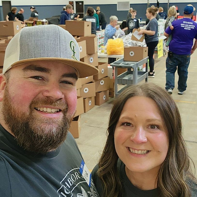 Two volunteers smiling at a community food packing event in a gymnasium.