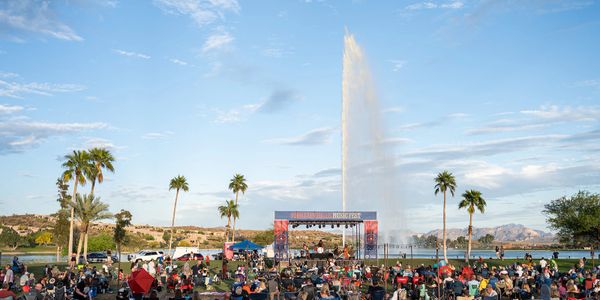 Outdoor concert by a lake with a large fountain and palm trees under a clear sky.
