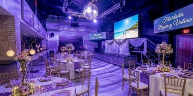 Elegant wedding reception with decorated tables and a dance floor under purple lighting.