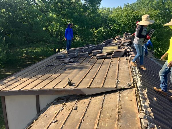 Roofers removing tiles from a house roof surrounded by trees on a sunny day.