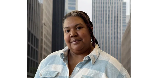 Woman in a blue and white checkered shirt with tall city buildings behind her.