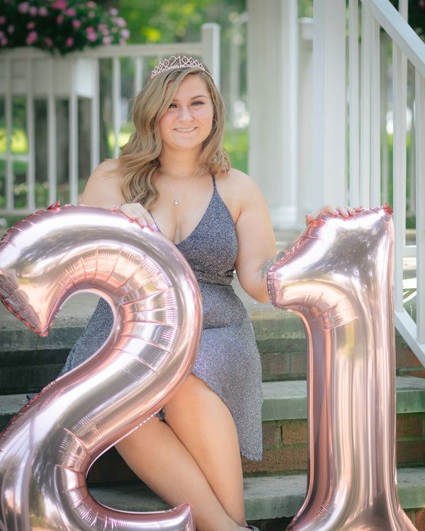 Young woman in a glittery dress and tiara, sitting on steps with large rose gold "21" balloons.

