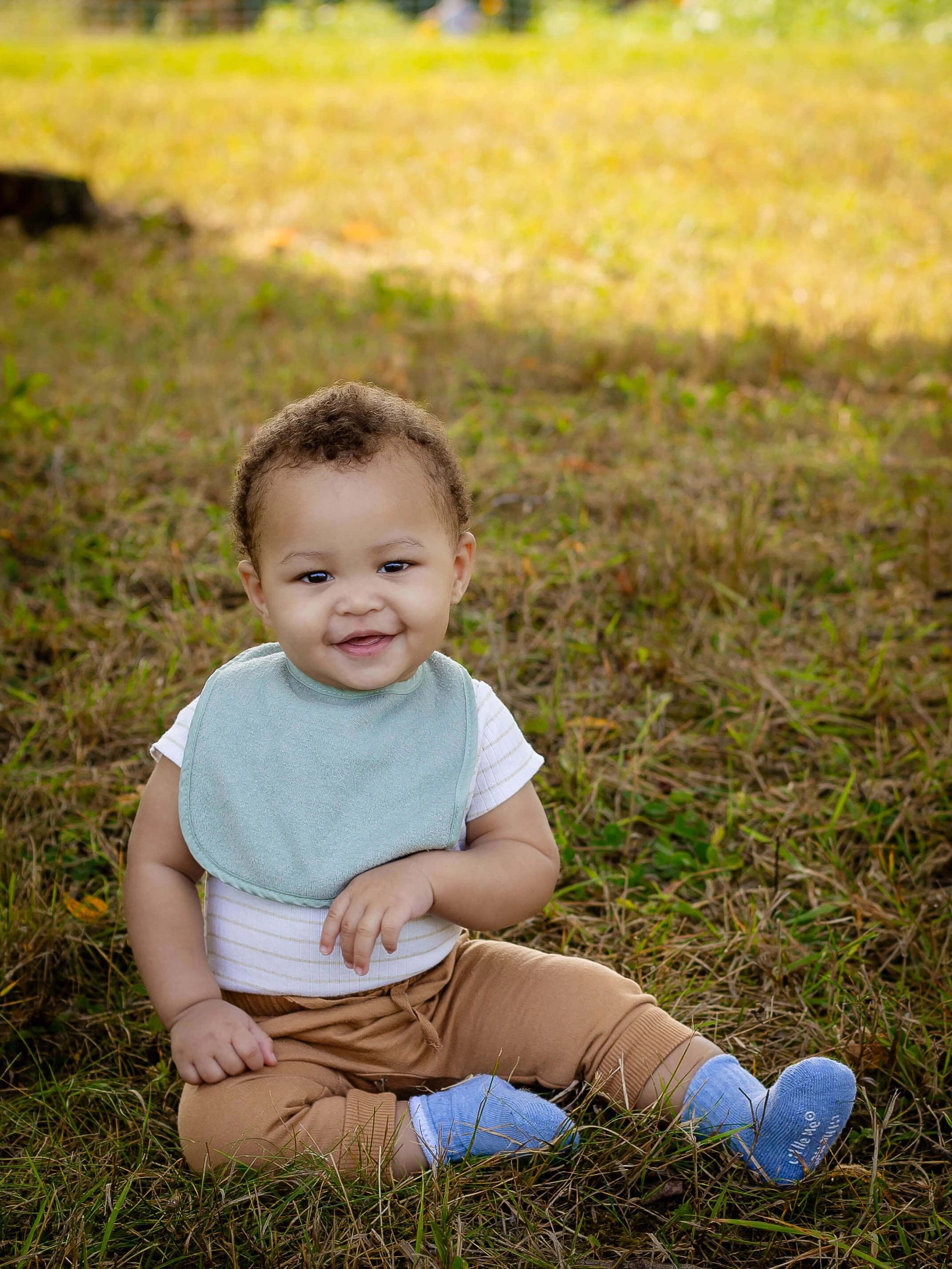 Baby sitting on grass wearing a striped shirt, tan pants, blue socks, and a light green bib, smiling