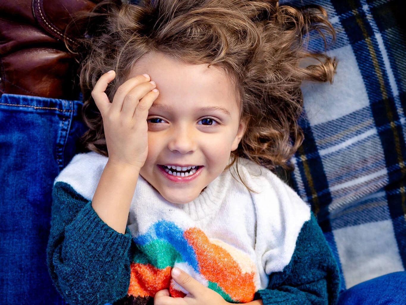 Smiling child with curly hair in a rainbow sweater, playfully covering one eye.