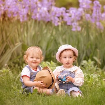 Two toddlers sitting on grass with flowers behind.