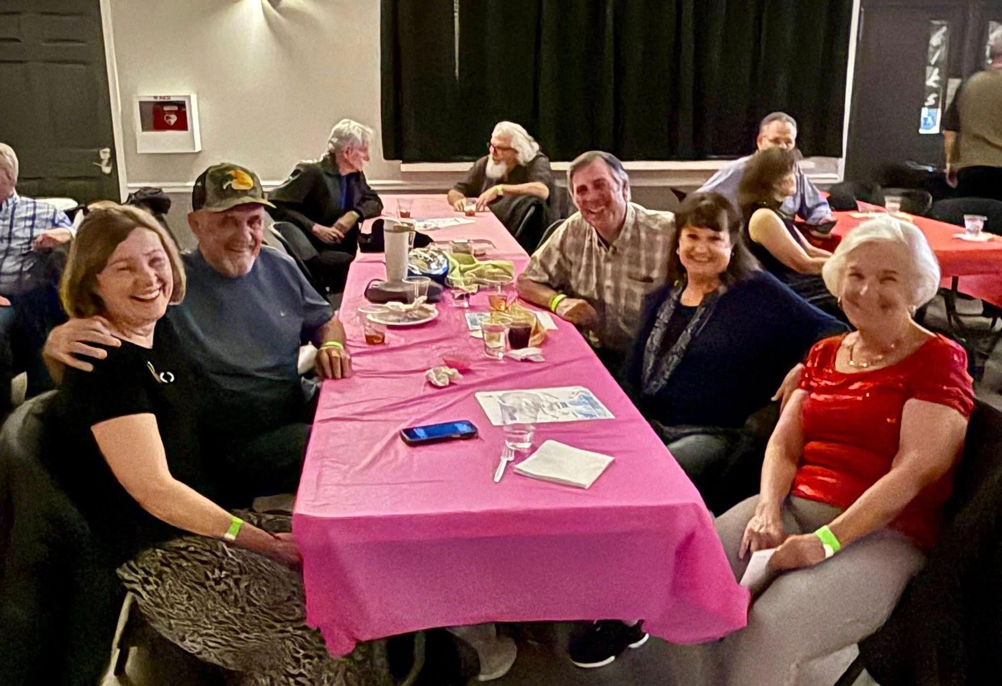 Group of smiling adults sitting around a table with pink tablecloth indoors.