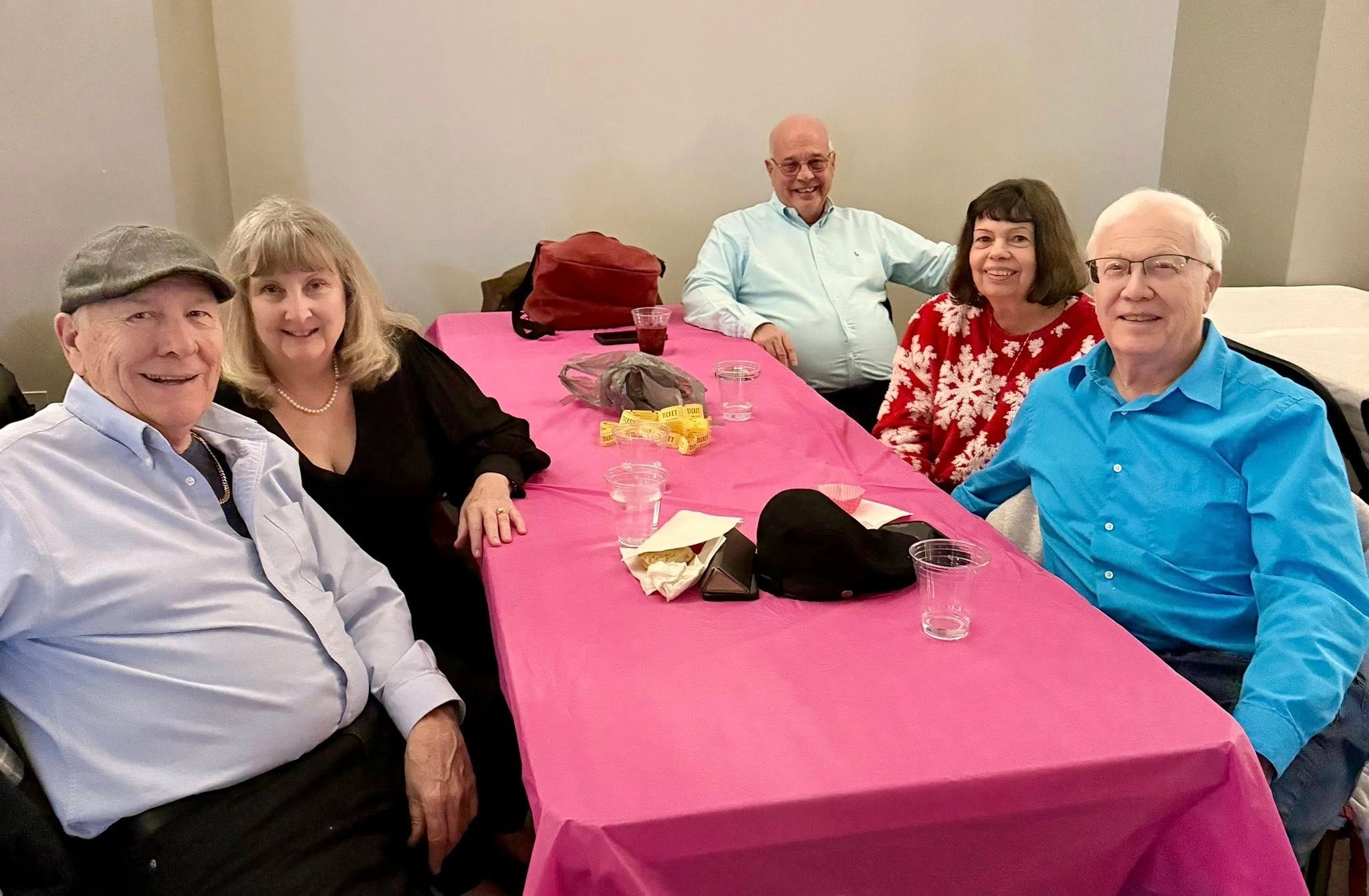Five elderly friends smiling around a pink table at a social gathering.