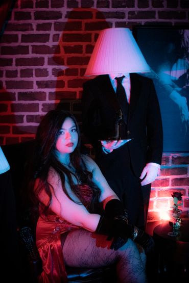 Woman in red dress seated with a person having a lampshade as a head in dim lighting.