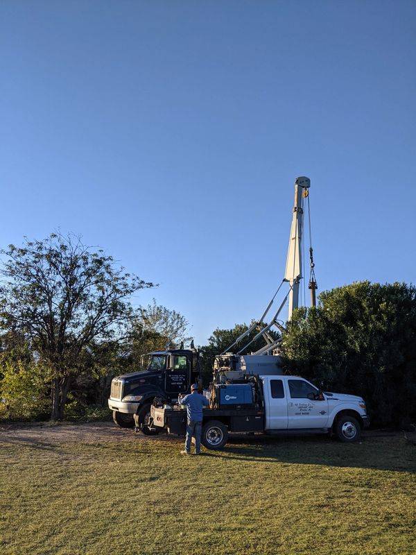 A man works near two trucks and a tall drilling rig in a grassy area under clear blue sky.