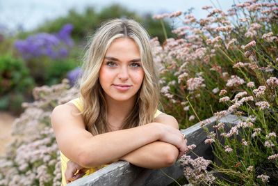 High school senior - young woman with blonde hair leaning on a wooden fence surrounded by flowers. 