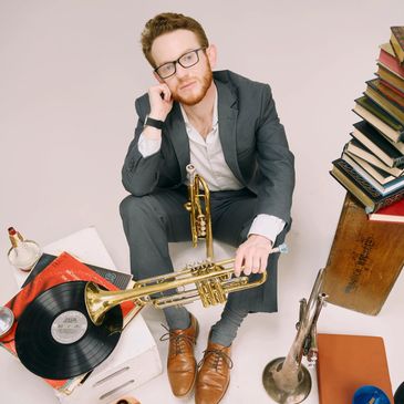 Man wearing glasses holding a trumpet and sitting surrounded by books and records
