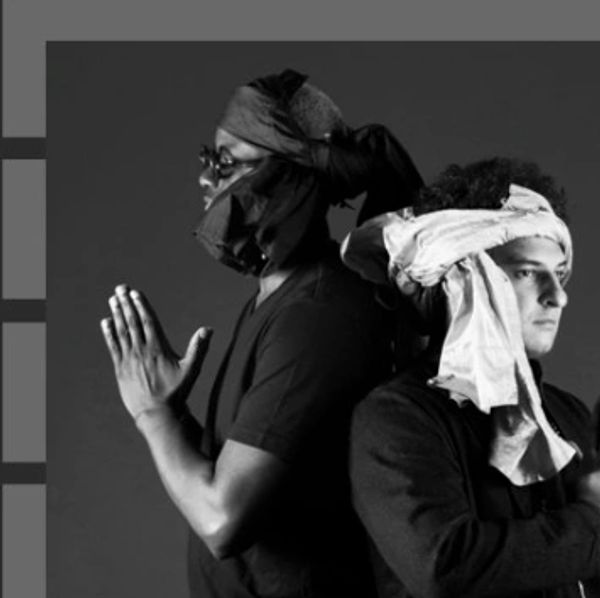 Two men standing back to back, one praying, both wearing head wraps.