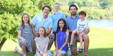 A smiling group of two men and five children posing outdoors on a sunny day.