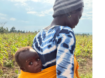 A woman carrying a baby on her back wrapped in an orange cloth in a field.