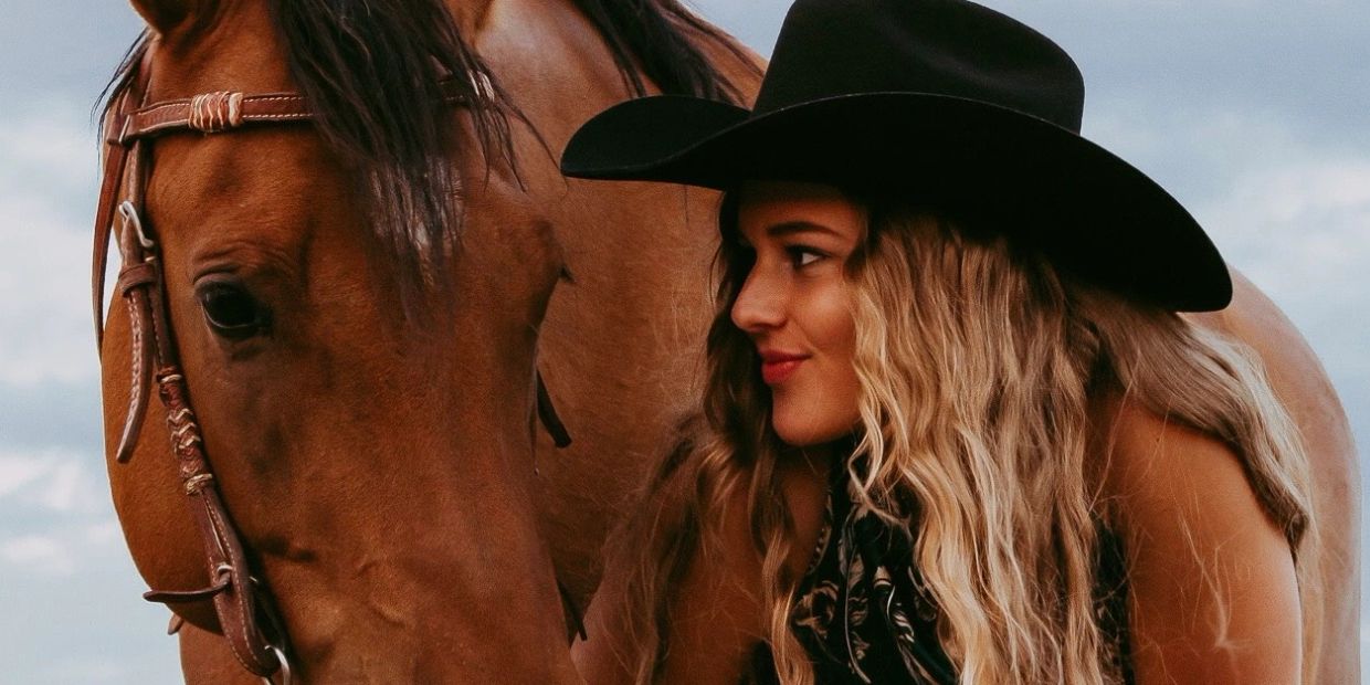A woman in a black hat smiles at a brown horse close by.