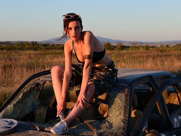 Young woman in camo pants sitting on a rusted car in a field at sunset.