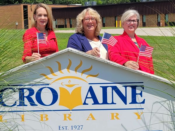 Three women holding American flags behind a library sign.