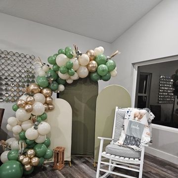 Green, white, and gold balloon arch with a cozy rocking chair and wooden lanterns in a modern room.