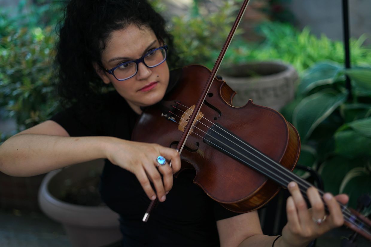 A light-skinned woman with black curly hair, playing viola, wearing a blue ring, in front of plants.