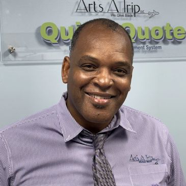 Smiling man in a checkered shirt and tie stands in front of a company sign.