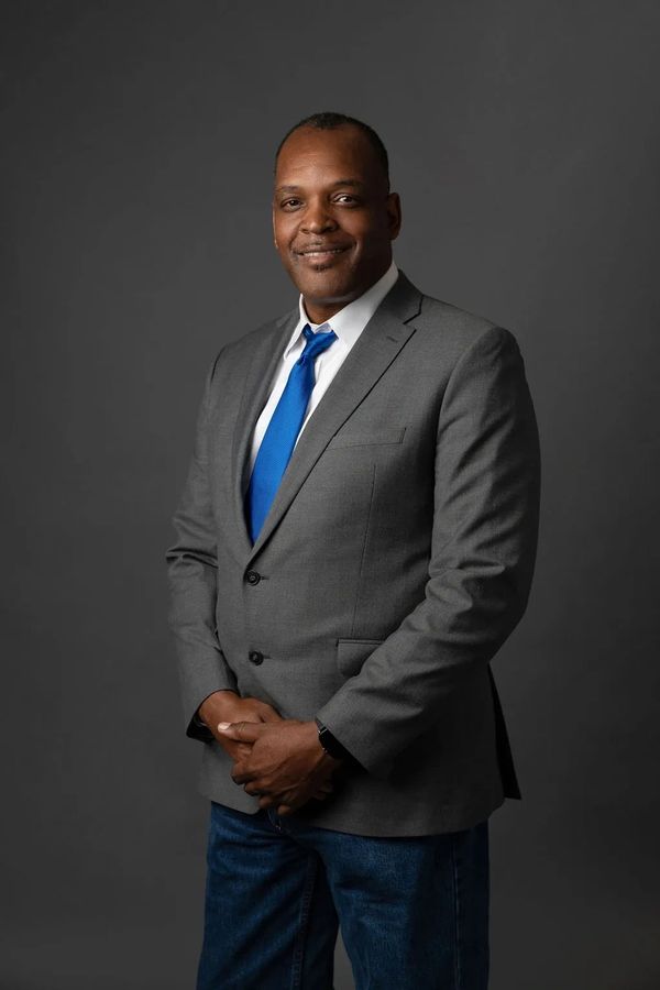 Man in a gray blazer with a blue tie smiles confidently against a dark background.