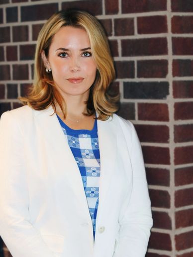 A woman with blonde hair wearing a white blazer and blue patterned top stands against a brick wall.
