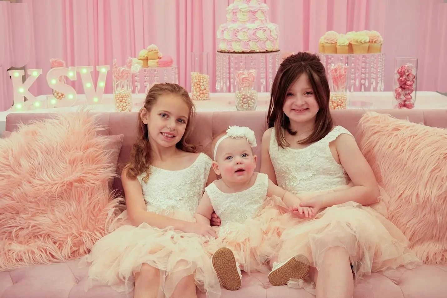 Three girls in matching dresses sitting on a pink couch at a pastel-themed party.