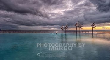 Storm clouds gathering at sunrise over the Cairns seaside pool and metal fish sculpture.