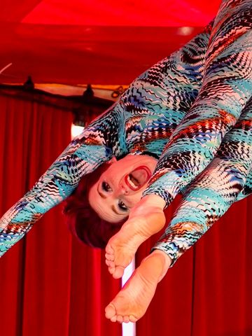 A contortionist in a colorful bodysuit performs an upside-down pose under red stage lights.