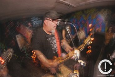 Blurry picture of a man with a grey goatee playing guitar and singing into a microphone. He is weari